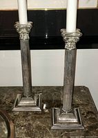 Photo showing both silver-plated Corinthian column candlesticks side by side on a marble tabletop, each holding a white candle.