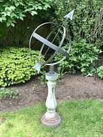 Full view of armillary sundial on stone pedestal in garden surrounded by plants and grass.