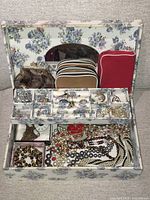 Floral fabric covered jewellery box open showing collection of costume jewellery organized in compartments and multiple small bags including earrings, rings, necklaces and bracelets.