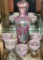 Full view of pink and lavender hand painted glass decanter with six matching glasses gathered around it on glass shelf.