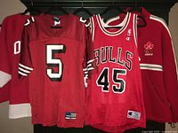 Two red jerseys hanging side by side on black rail, one football and one basketball jersey seen in context