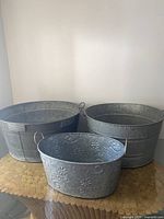 Photo showing three galvanized steel tubs: two large ones in the background and a smaller decorative one in front on a wooden surface.