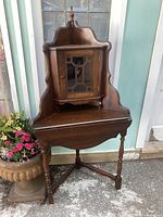 Front view of antique corner cabinet showing door with glass panel and wooden lattice, shaped back with finial, and drop-leaf table raised on turned legs.