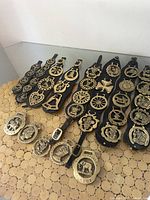 Eight sets of horse and pony brass ornaments attached to black leather straps and loose brass pieces shown on round wooden placemat surface under bright lighting.