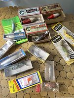 Photo showing an assortment of vintage fishing lures, some in original boxes with advertisements, others in plastic cases, and some in original card packaging.