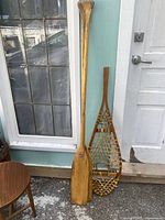 Photo showing two wooden paddles standing against wall next to a traditional snowshoe with woven netting.
