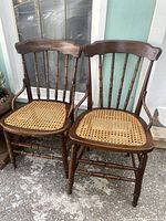 Pair of antique wooden gun stock chairs with cane seats, shown side by side on a porch.