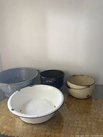 Four vintage enamelled and cast iron pots with varied colors and sizes showing wear, including cream and green enamel, black and white GSW, blue and white enamel tub, and cast iron caldron.