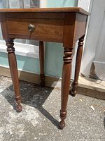 Front view of a square vintage solid wood side table with a single drawer and round brass knob. Turned legs with decorative details. The bottom part of the legs are visible with turned design.