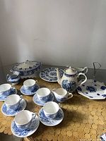 Full set including cups, saucers, teapot, serving plates, gravy boat, creamer, and lidded dish displayed on textured table covering with blue and white china patterns visible