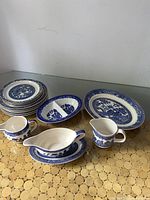 Full lot view showing multiple Blue Willow patterned plates, divided dish, oval bowl, creamers, gravy boat, and underplate on round table.