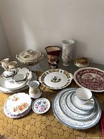 Full lot of vintage china items including plates, vase, lidded dish, mug, and figurine on a table with gold patterned cloth