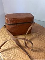 Brown textured leather carrying case with zipper and long strap, closed and resting on a wooden surface.