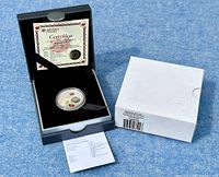Black presentation box containing the silver proof coin, certificate of authenticity, and a white outer box to the side on a blue textured surface.
