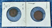 Two coins in white cardboard holders with circular windows, showing front and back sides of the coins.