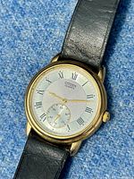 Close-up image of the Citizen men's dress watch face showing white dial with black Roman numerals, sub-second dial, gold-tone casing, and black leather strap.