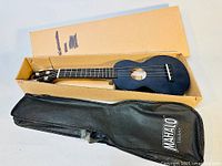 Black soprano ukulele in original cardboard box with a black gig bag beside it.
