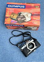 Digital camera body with front view, showing black leather case and strap, positioned in front of original color box with product details.