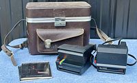 Polaroid Spirit and Sun 600 cameras alongside brown leather carrying case and instruction manual, showing full lot contents