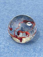 Clear round glass paperweight with internal red swirls on blue snow background, top view.