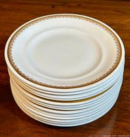 Stack of 13 white fine bone china plates with a gold floral border on rim, on wooden surface.