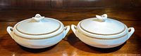 Two white Copeland-Spode bone china lidded casserole dishes with gold decorative trim, displayed side by side on a wooden surface.