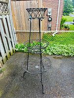 Full view of the 3-tier black metal plant stand showing its height and overall structure outdoors on pavement near a chain-link fence.