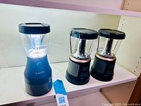 Photo of 3 portable LED lanterns on white shelf: one blue Duracell Hybrid model and two smaller black and gray lanterns.