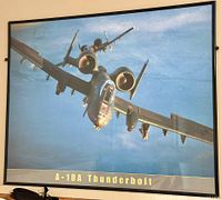 Framed photo showing A-10A Thunderbolt jet in mid-flight with blue sky background, close-up with black frame and text beneath image.