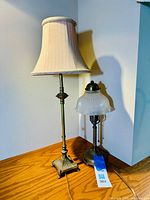 Both lamps displayed on wood surface; shorter antique style lamp has a white ribbed ruffled glass shade with brass base, taller lamp has beige fabric shade and square ornate base.