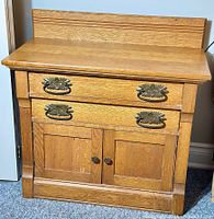 Front view of antique oak washstand showing two drawers with decorative metal handles above a two-door cabinet, natural wood finish.
