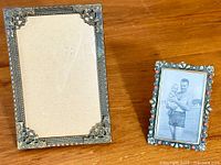 Two antique picture frames on wood surface, larger empty ornate metal one and smaller one with coral cabochons and photo.