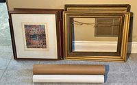 Photo showing two smaller size framed McMichael Canadian Collection Group of Seven prints depicting autumn trees and water, one in brown wood frame with matting and another smaller print with a similar frame. Next to prints are two empty frames, one gold-toned and one brown wood. Two rolled posters in tubes lie in front.