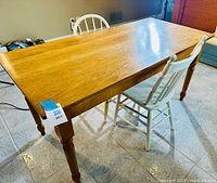 Pine rectangular dining table with turned legs and a drawer, accompanied by two white painted wooden spindle-back chairs, visible on tiled floor.