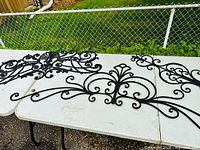 Three black ornate wrought metal wall hangings displayed on white folding tables outdoors with grass and chain link fence in background. Each piece features intricate scrollwork and decorative motifs.