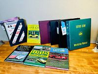 Photo showing a collection of books and binders including a green Lotus Elan Workshop Manual and several Lotus books and periodicals.
