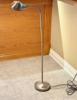 Full view of chrome gooseneck floor lamp standing on tiled floor against wood paneling and cream wall showing overall structure and base with small dent.