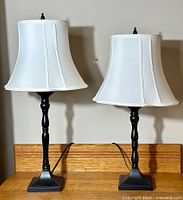 Pair of black metal table lamps on wooden surface with white fabric shades, one lamp taller than the other