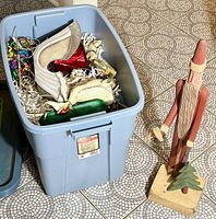 Photo showing the open Rubbermaid bin filled with various Christmas decorations placed on floor with tall wooden Santa Claus figurine standing next to it.