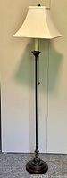 Full view of traditional style floor lamp showing dark metal stand and white fabric shade, pull chain switch visible, lamp standing upright on carpeted floor with plain wall background.