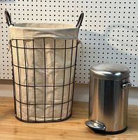 Photo showing the black metal wire laundry hamper with beige canvas liner alongside the stainless steel pedal garbage can side by side on wooden surface.