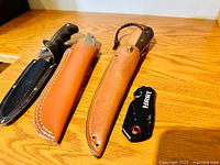 Four cutting utensils displayed on wooden surface: two fixed-blade knives with sheaths, one folding knife, one cutting tool.