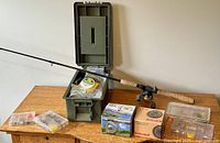 Fishing rod with attached reel, green tackle box and multiple small cases and reel boxes arranged on a wooden surface.