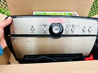 Front view of FoodSaver vacuum sealing machine inside box with partial roll and full box of bags behind
