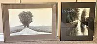 Two framed black and white photographic prints placed side by side on a carpeted floor; left print showing a rural road lined with trees, right print shows Edward Weston's reflective water scene.