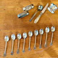 Full view of all items including napkin rings, demitasse spoon, antique napkin ring, cheese cutter, and silver plated teaspoons.