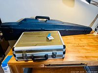 Two hardshell cases displayed on wooden surface: large blue plastic case in back, silver aluminum case with black handle in front.