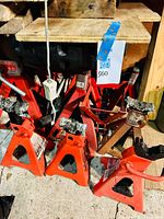 Several orange and red car jack stands positioned on a concrete floor beneath wooden boards, including some with visible rust and wear.