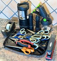Tray holding scissors, stapler, tape dispenser, flashlights, and small tools arranged on granite countertop with tile background.