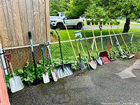 Wide shot showing a lineup of various garden shovels, spades, rakes, and pruners all leaning against a chain link fence outdoors on a pavement area